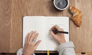 woman writing in notebook, cup of coffee and crossaint nearby